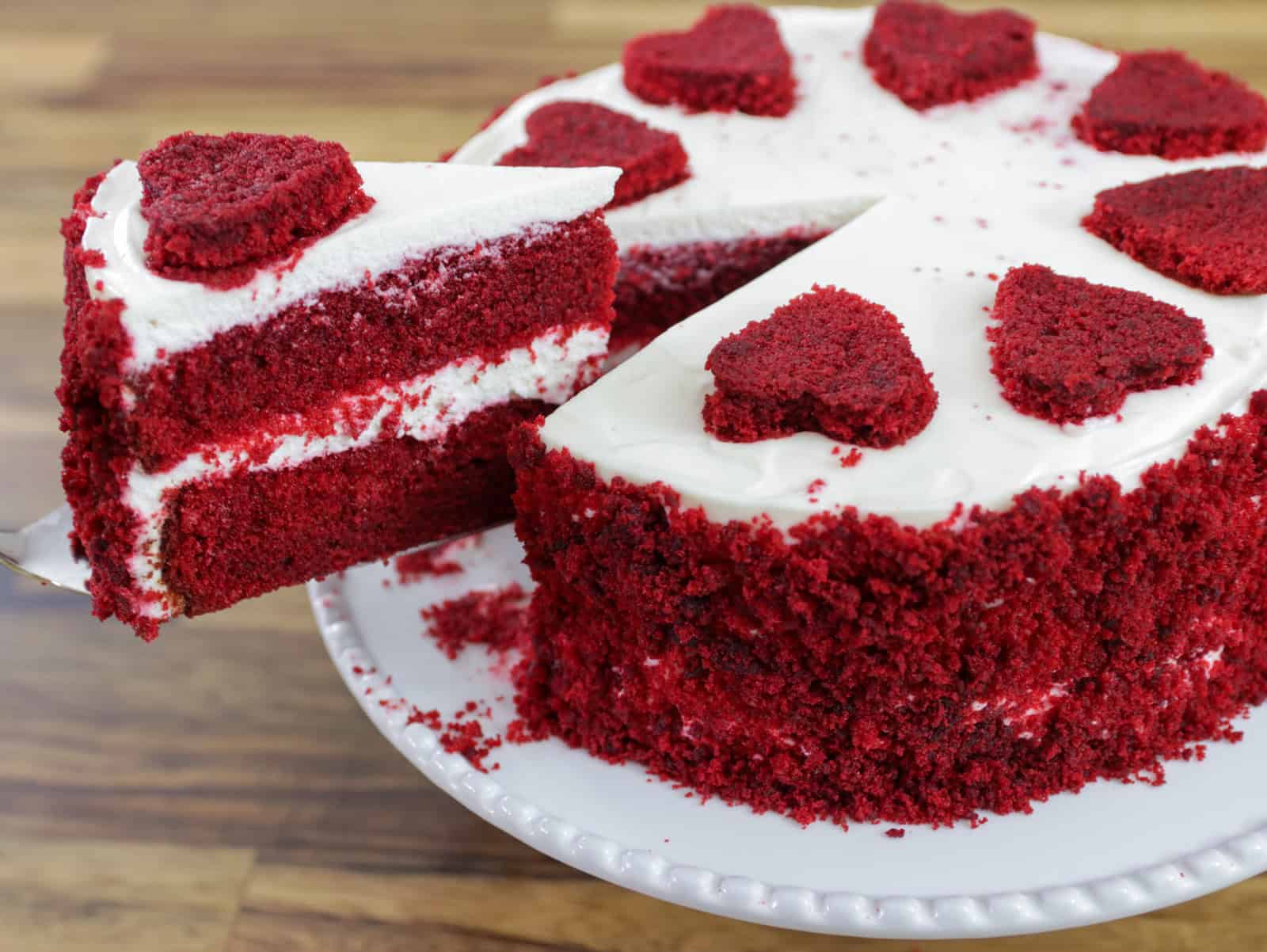 A slice of red velvet cake with white frosting is being lifted from a round cake decorated with heart-shaped pieces on top.