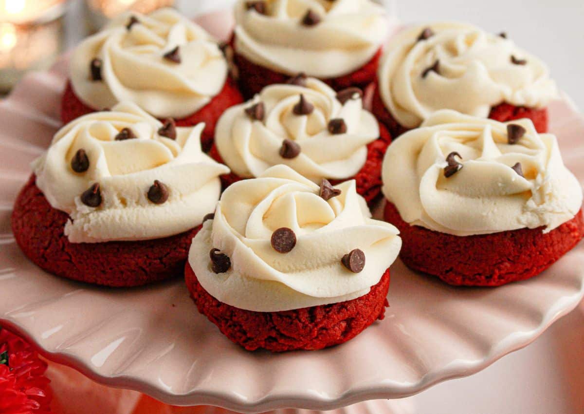 Red velvet cookies topped with swirls of white frosting and decorated with chocolate chips, arranged on a pink cake stand.