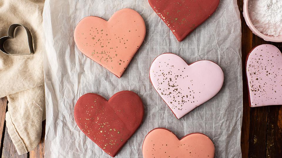 Five heart-shaped cookies decorated with pink and red icing and gold sprinkles are arranged on parchment paper, with a heart-shaped cookie cutter and a bowl of powdered sugar nearby.