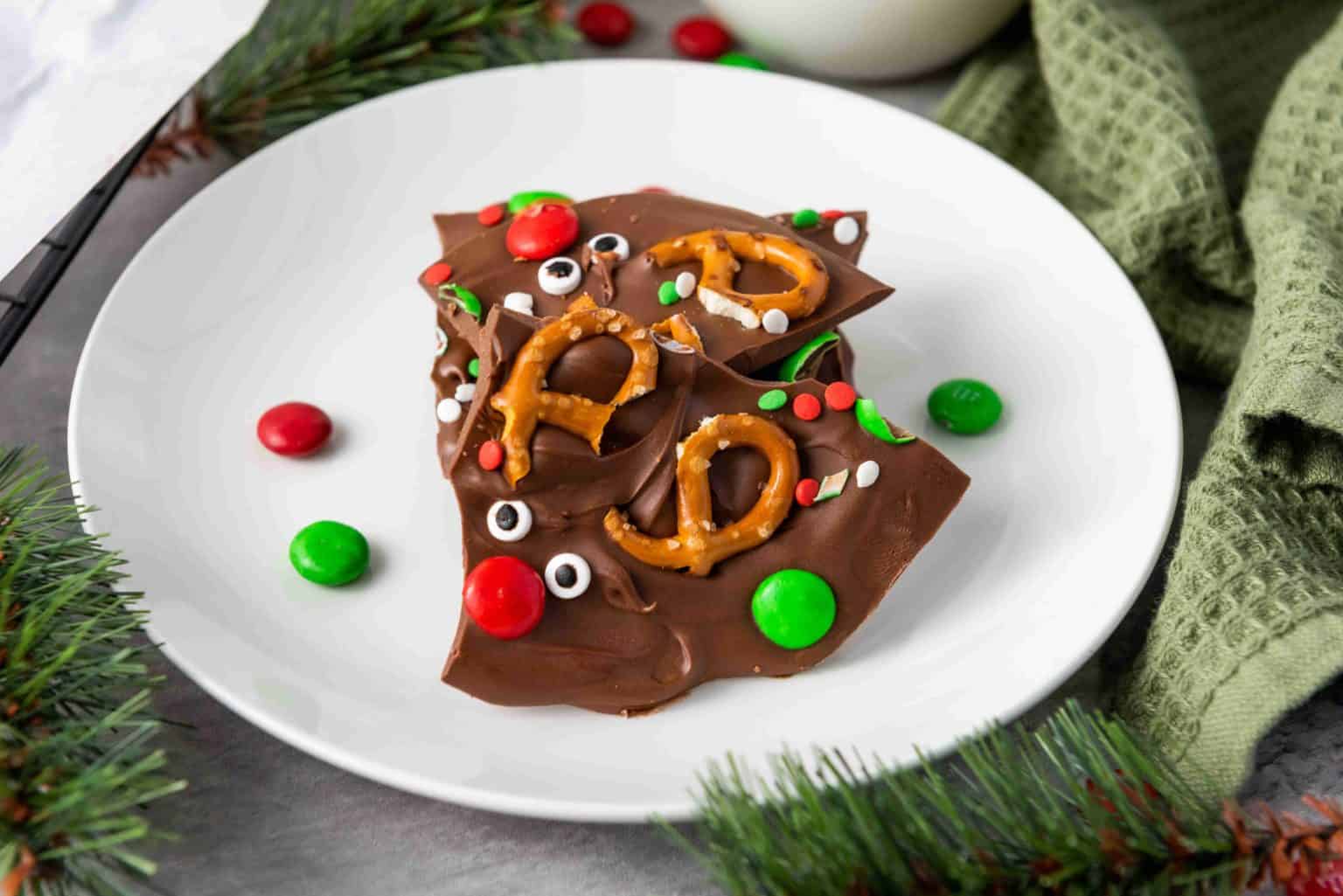 Three pieces of chocolate bark topped with pretzels, red and green candy-coated chocolates, and sprinkles on a white plate.