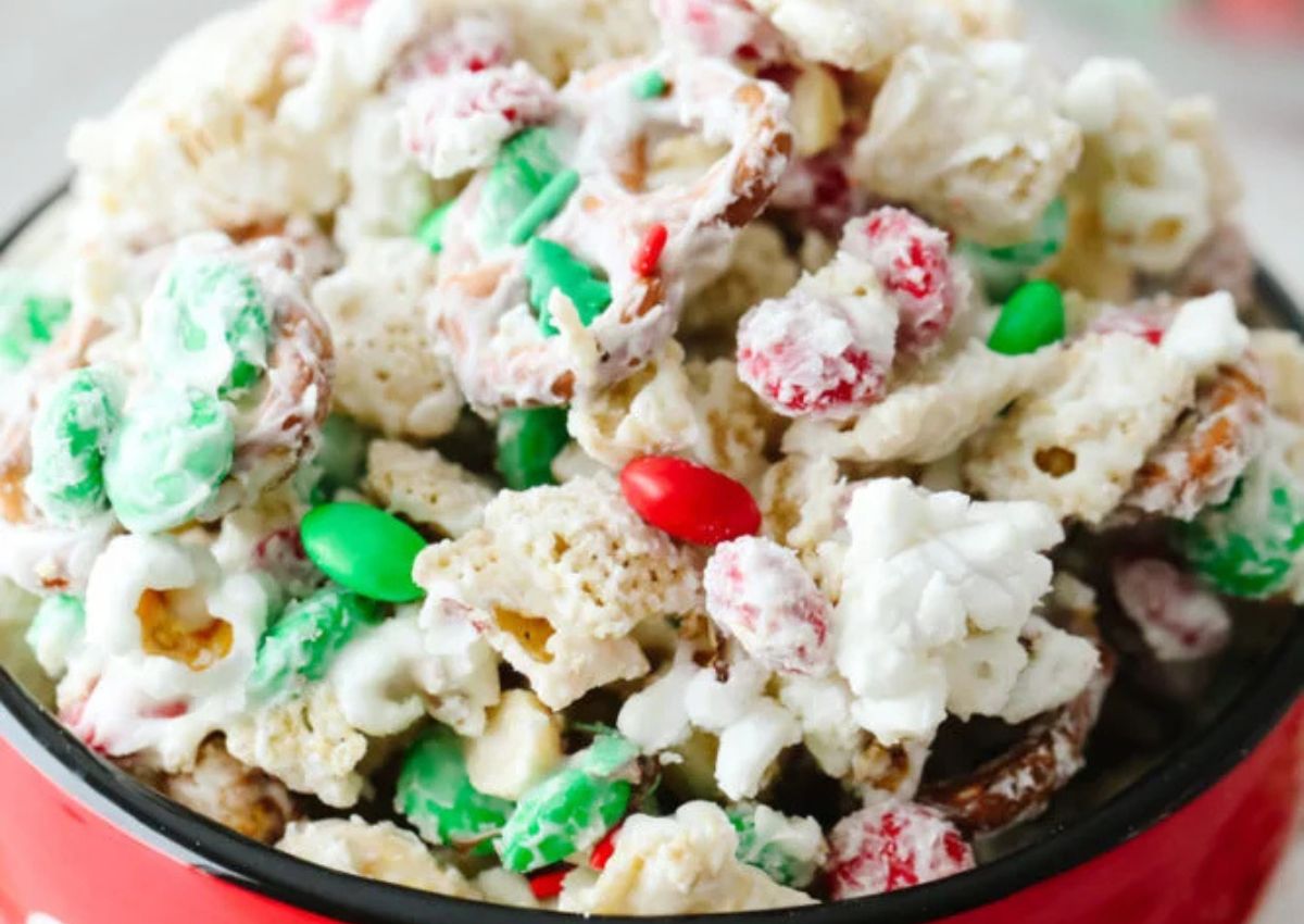 A bowl filled with a snack mix of popcorn, pretzels, cereal, and red and green candy pieces, all coated in white chocolate.