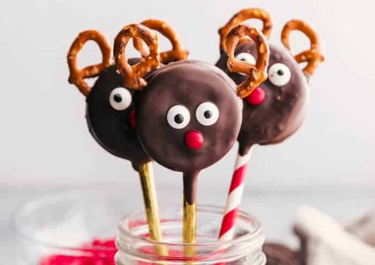 Three chocolate-covered cookie pops decorated as reindeer with candy eyes, red noses, and pretzel antlers, placed in a glass jar.
