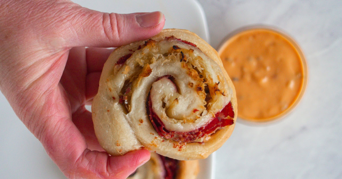 A hand holding a pinwheel-shaped pastry filled with cheese and meat, with a cup of orange dipping sauce in the background.