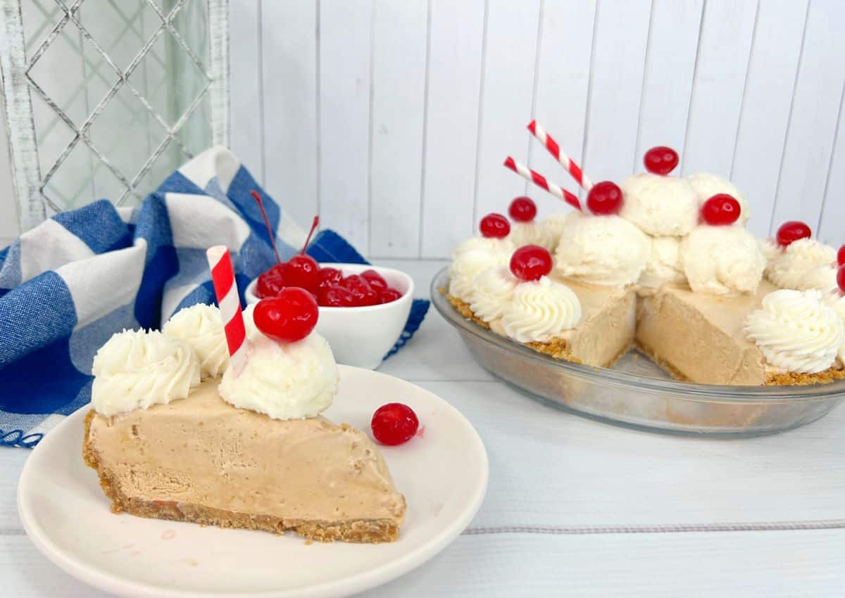 Slice of brown ice cream pie topped with whipped cream and cherries on a plate, with the remaining pie and a bowl of cherries in the background.