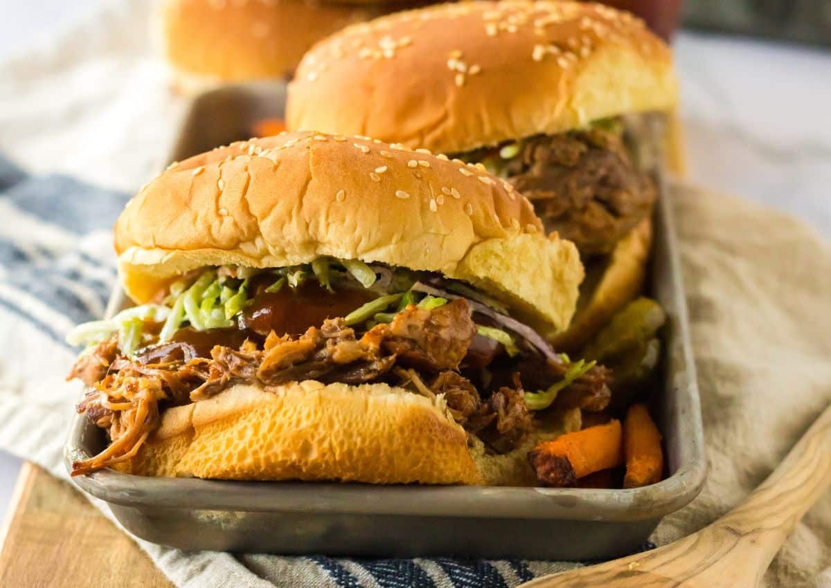 Two pulled pork sandwiches on sesame buns, topped with shredded vegetables, served on a tray with pickles and fries.