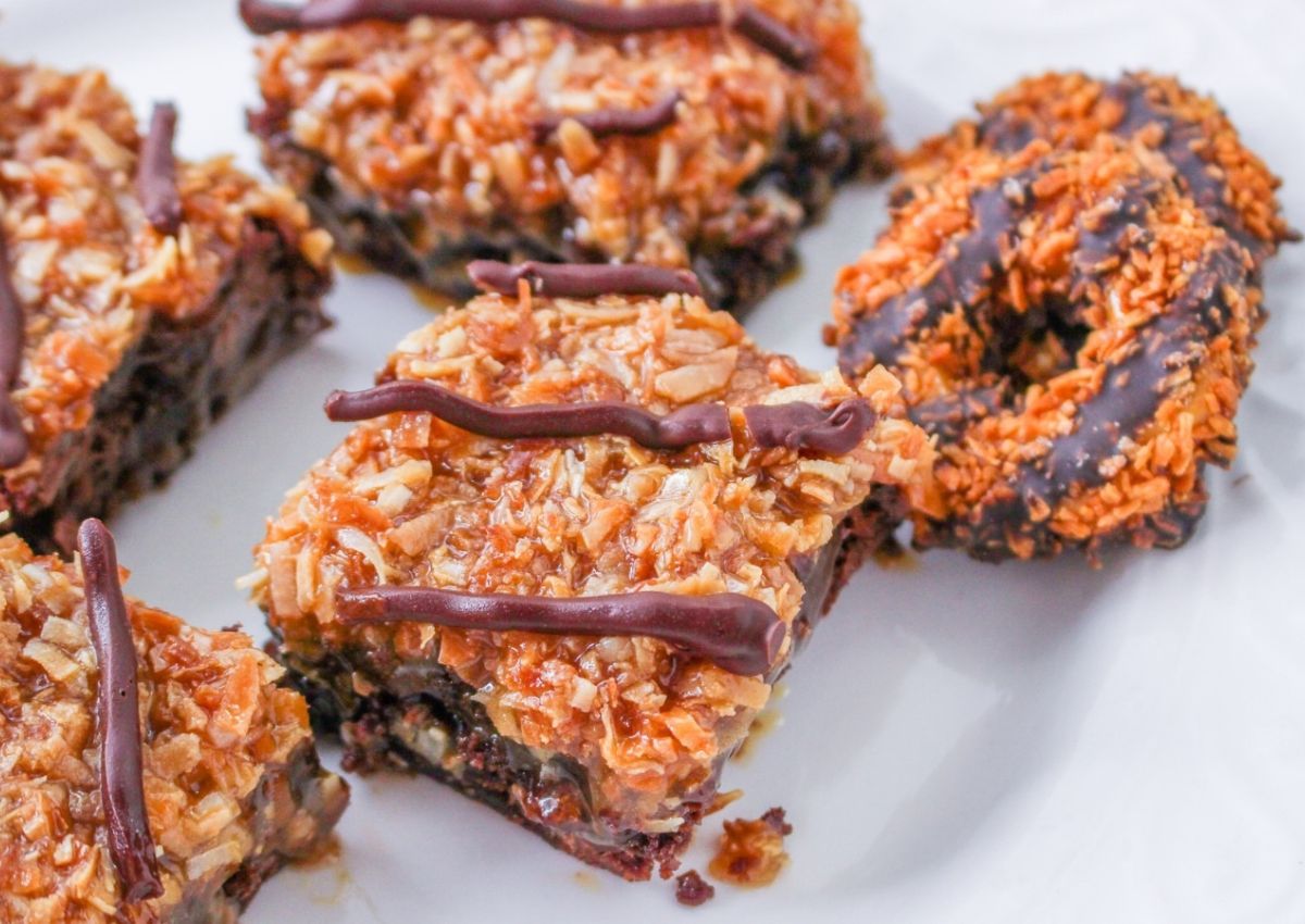 Close-up of dessert bars and cookies with a coconut and caramel topping, drizzled with chocolate, on a white plate.