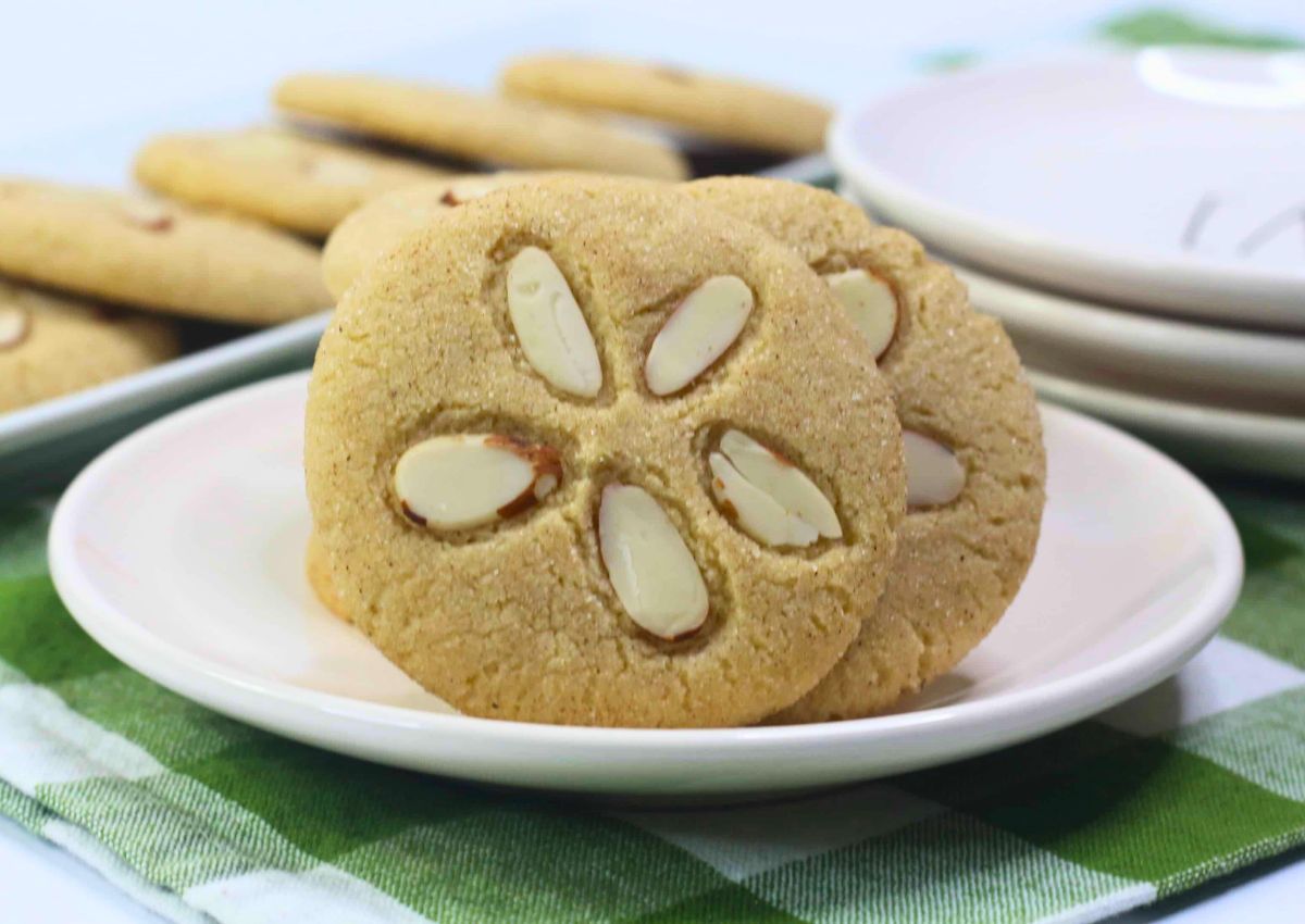 A plate of round cookies topped with sliced almonds, placed on a green and white checkered cloth, with more cookies and stacked plates in the background.