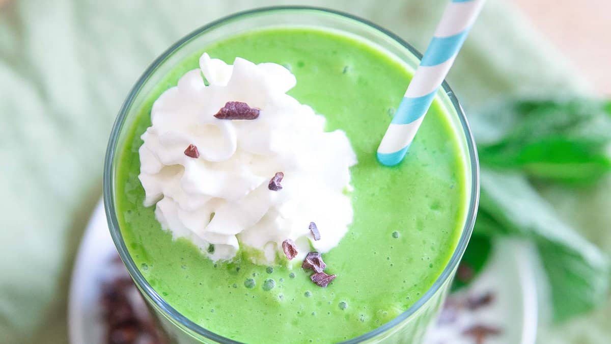 A green smoothie topped with whipped cream and chocolate shavings, served with a blue-striped straw.