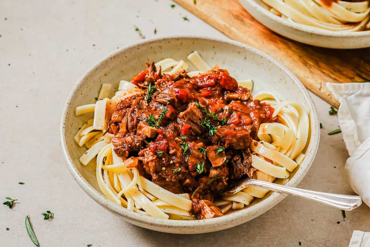 A bowl of fettuccine pasta topped with a chunky tomato-based meat sauce, garnished with fresh herbs, with a fork resting in the bowl.