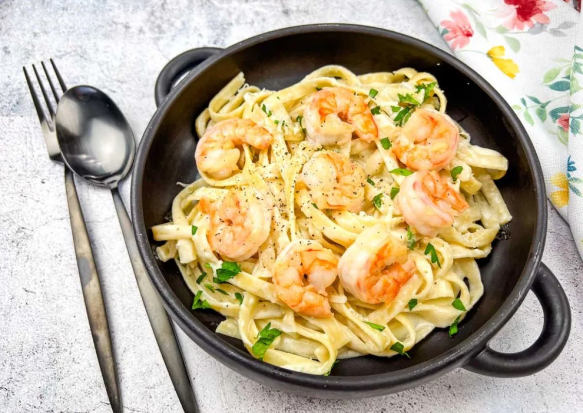 A black bowl filled with shrimp fettuccine Alfredo, garnished with chopped parsley, sits beside a spoon and fork on a light textured surface.