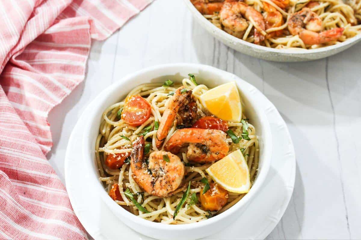 A bowl of spaghetti with shrimp, cherry tomatoes, herbs, and lemon wedges, placed on a white surface next to a red-striped cloth.