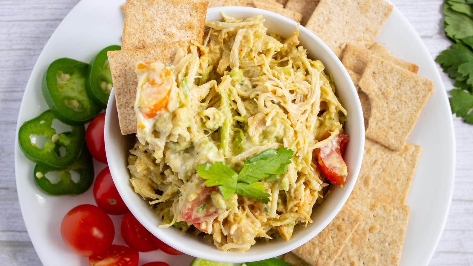 A bowl of shredded chicken salad garnished with parsley, surrounded by crackers, sliced cherry tomatoes, and jalapeรฑo slices on a white plate.