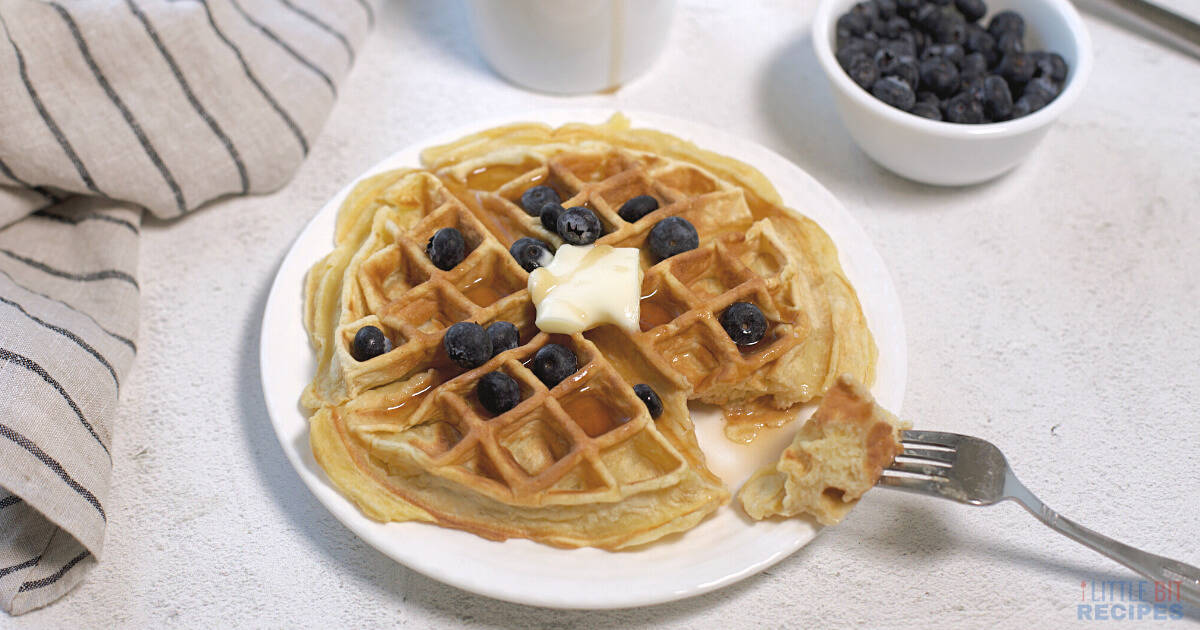 A plate with a waffle topped with butter, blueberries, and syrup; a fork with a waffle piece, a bowl of blueberries, a mug, and a striped napkin are nearby.