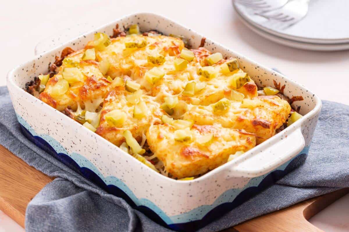 A baked casserole topped with melted cheese, diced pickles, and tater tots, placed in a rectangular ceramic dish on a wooden board.
