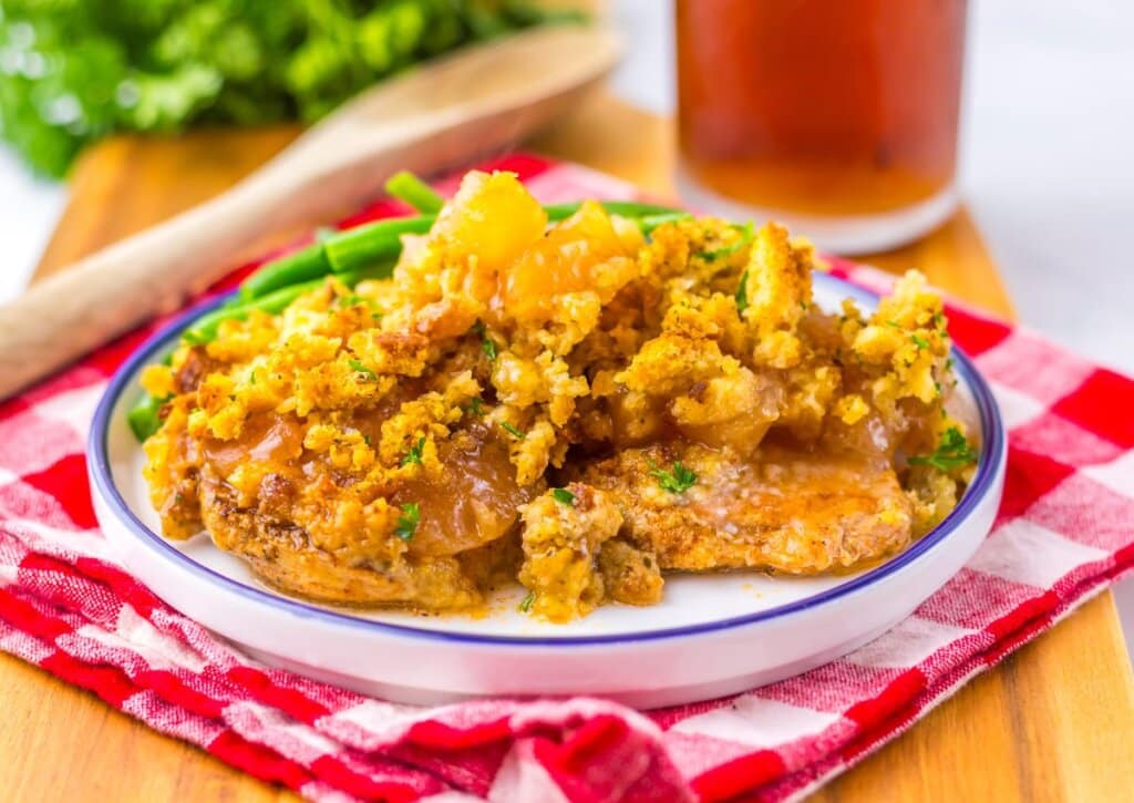 A plate of baked pork chops topped with a golden, crumbly stuffing, with green beans on the side, set on a red checkered cloth.