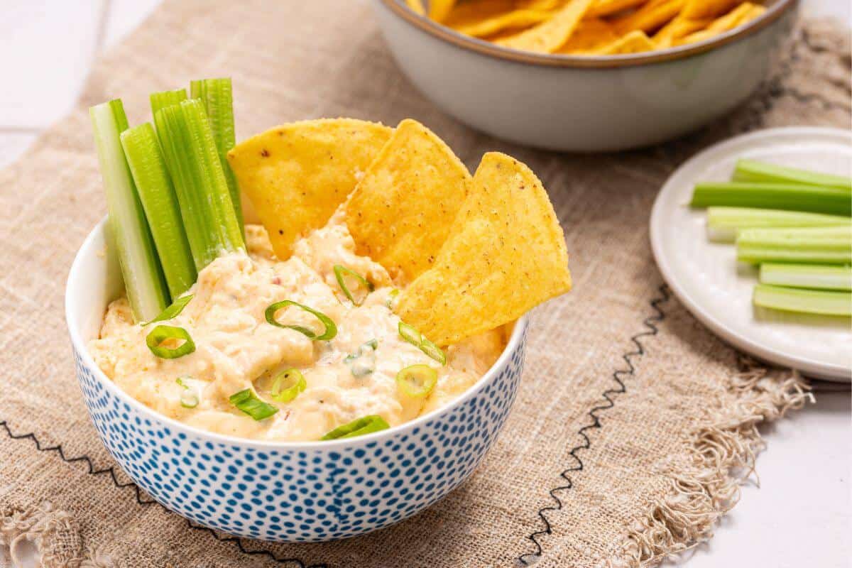 A bowl of creamy dip topped with sliced green onions, served with celery sticks and tortilla chips, sits on a textured cloth next to a plate of celery and a bowl of chips.