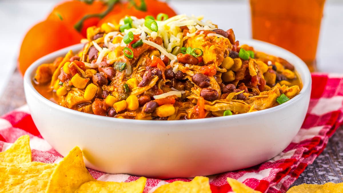 A bowl of chili with beans, corn, cheese, and green onions, served with tortilla chips on a red checkered cloth.