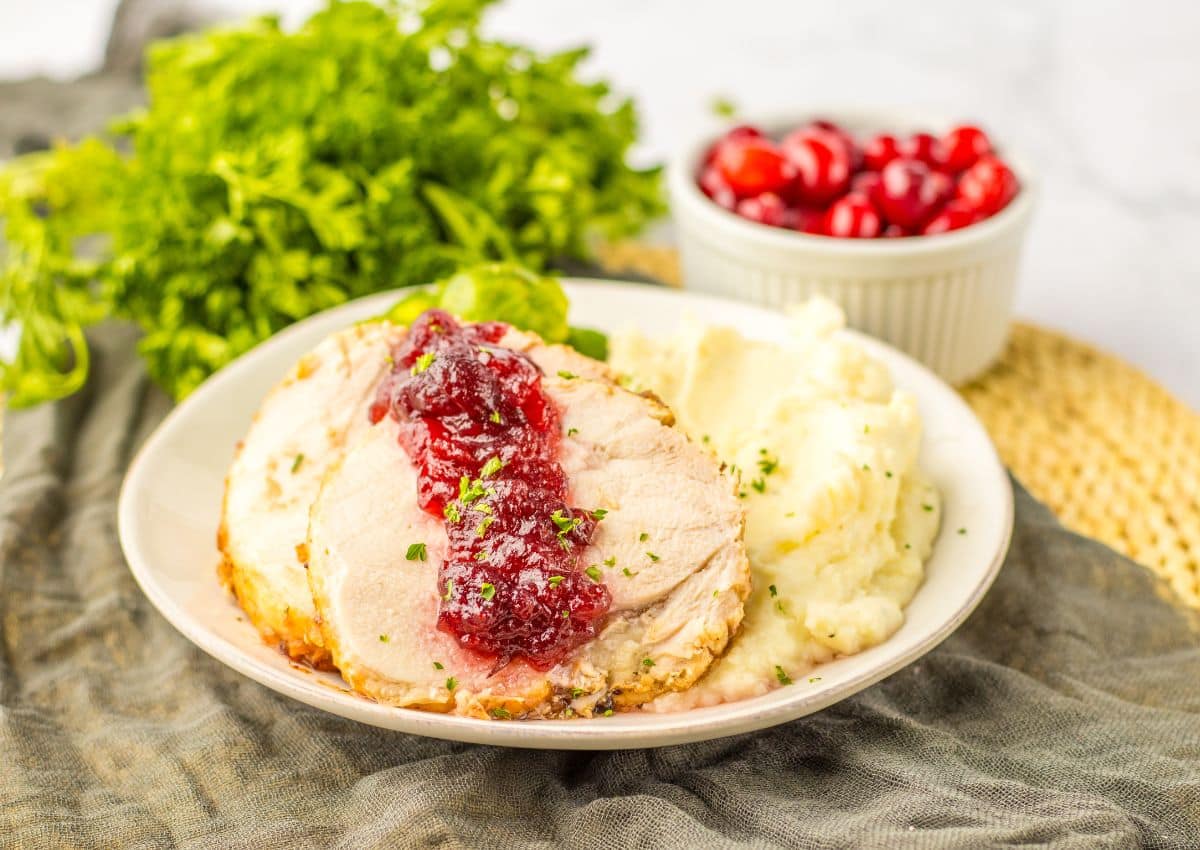 A plate with slices of turkey topped with cranberry sauce, served alongside mashed potatoes; a bowl of cranberries and a bunch of parsley are in the background.
