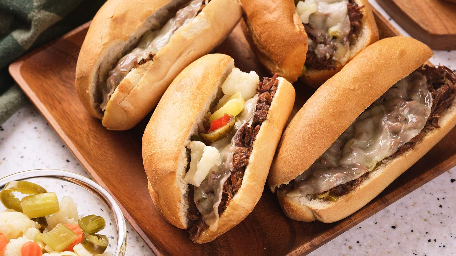 Four Italian beef sandwiches on a wooden tray, filled with shredded beef, melted cheese, and pickled vegetables. A small bowl of pickled vegetables sits nearby.