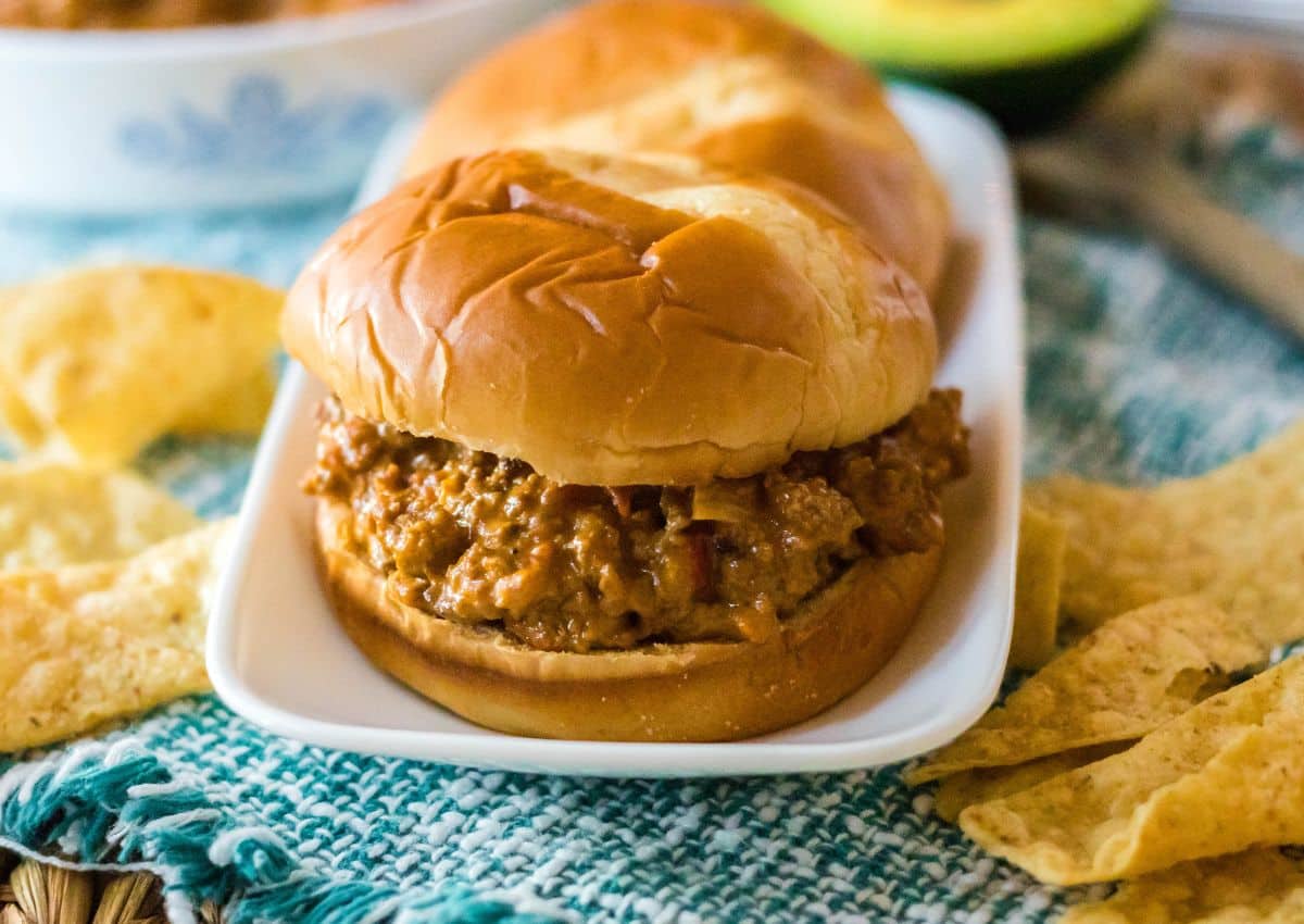 A sloppy joe sandwich with a meat filling on a bun sits on a white plate, surrounded by tortilla chips.