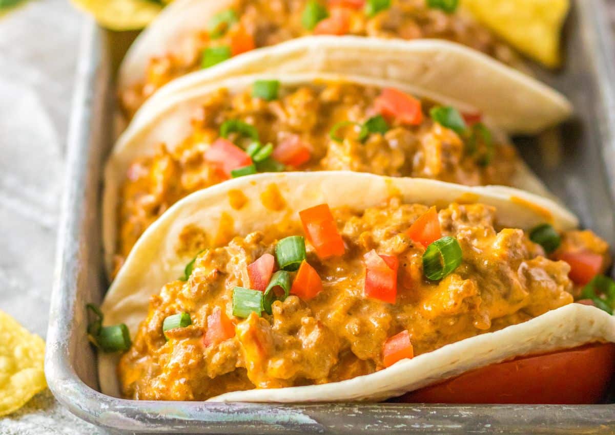 Soft tacos filled with a creamy ground beef mixture, topped with diced tomatoes and chopped green onions, served in a metal tray.