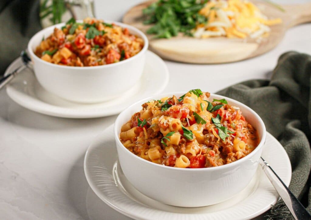Two white bowls filled with pasta in a tomato-based sauce, garnished with chopped herbs, sit on saucers with spoons; shredded cheese and herbs are in the background.