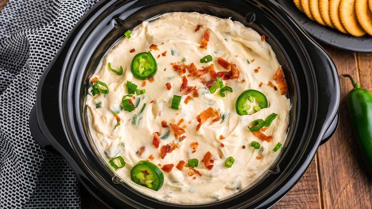 A slow cooker filled with creamy dip topped with bacon pieces, sliced jalapeรฑos, and chopped green onions, with crackers and a jalapeรฑo pepper nearby.