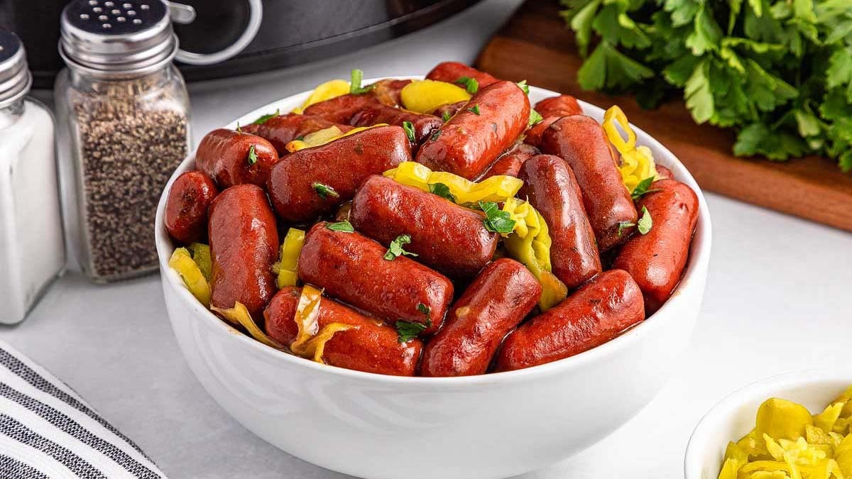 A white bowl filled with cocktail sausages and sliced yellow peppers, garnished with chopped parsley, sits on a countertop near salt and pepper shakers.