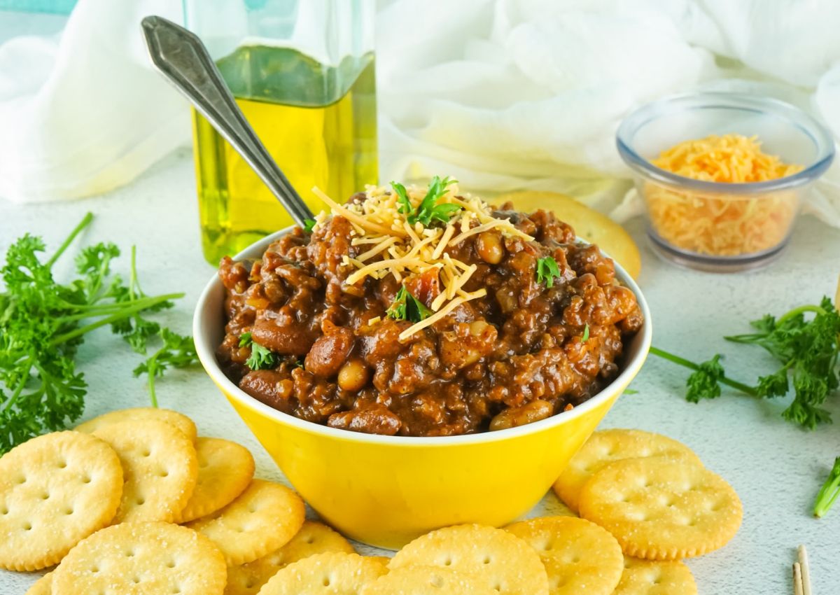 A bowl of chili topped with shredded cheese and parsley, surrounded by round crackers, with a bottle of oil and a bowl of shredded cheese in the background.