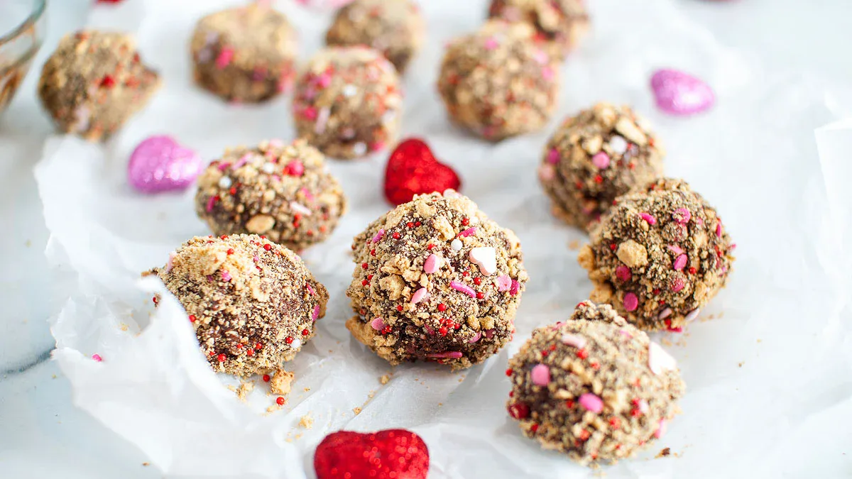 Chocolate truffles coated in crushed cookies and pink sprinkles are arranged on white parchment paper with red and pink heart decorations.
