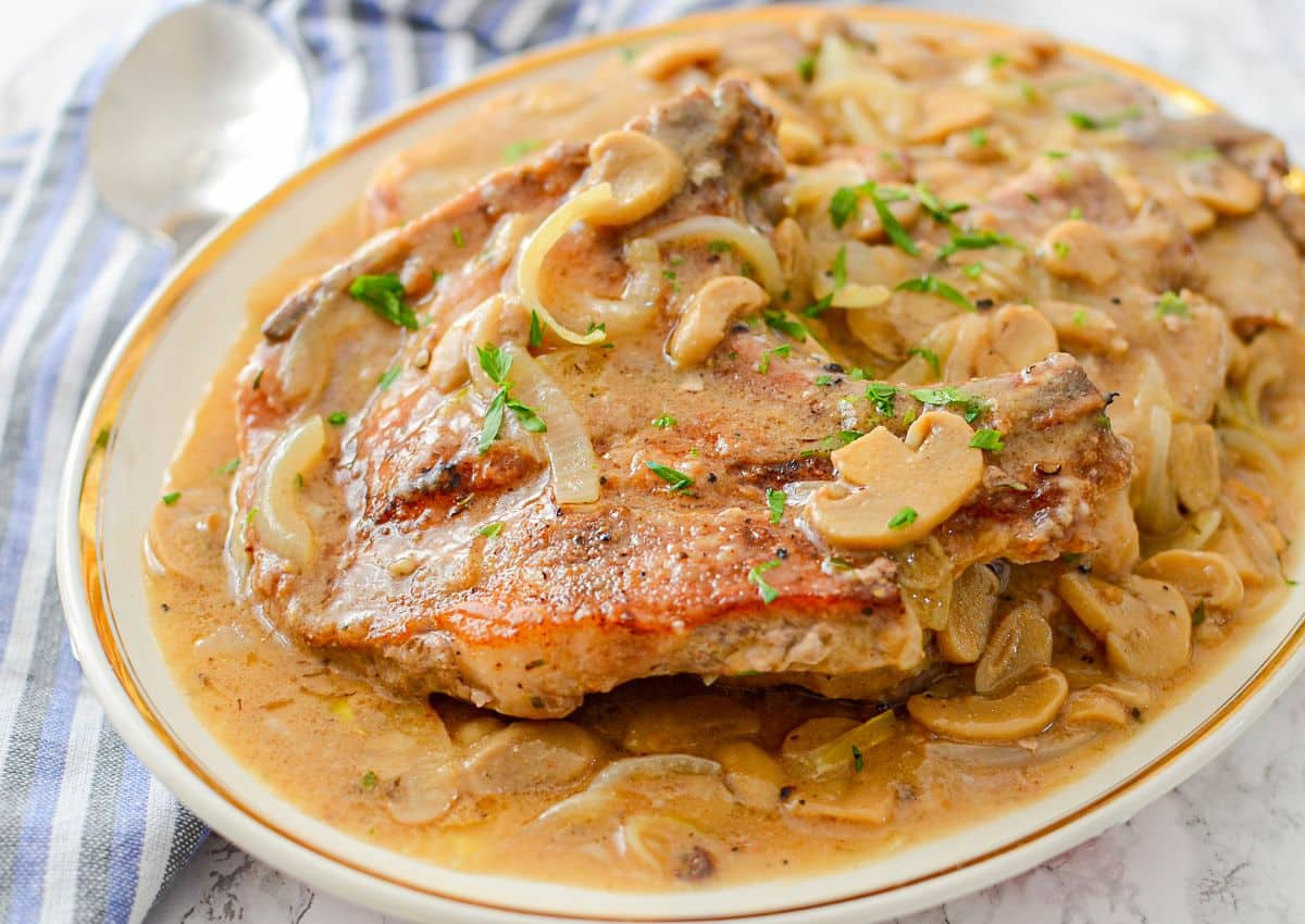 Two pork chops served on a plate, covered in creamy mushroom and onion sauce, garnished with chopped parsley. A spoon and striped cloth are in the background.