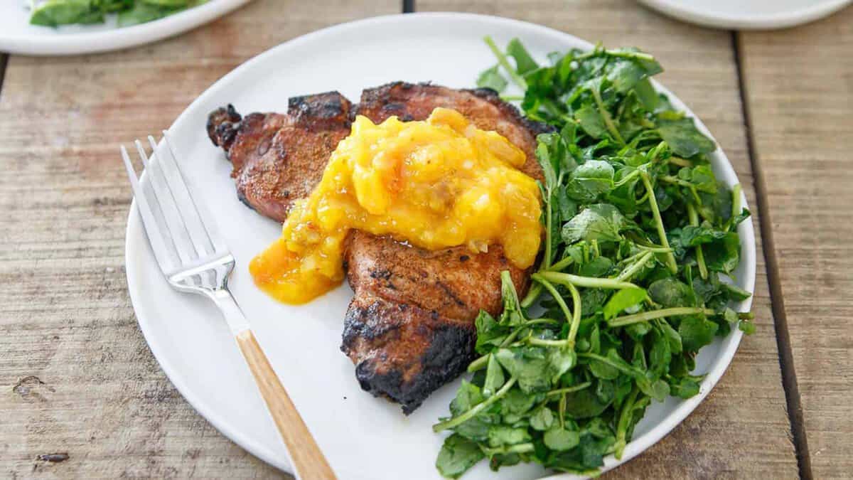 Grilled pork chop topped with a yellow fruit chutney, served with a side of fresh green salad on a white plate with a fork.