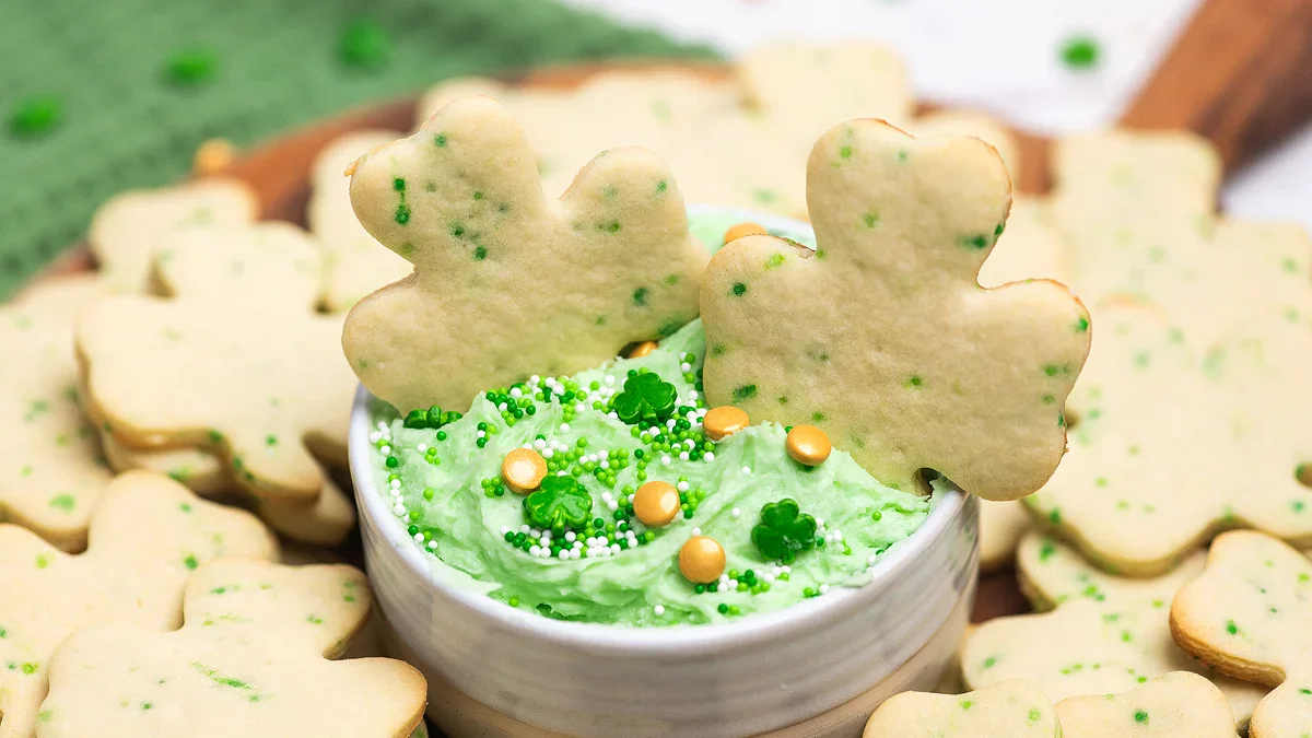 Shamrock-shaped sugar cookies with green frosting and sprinkles in a small bowl, surrounded by more cookies on a wooden board.