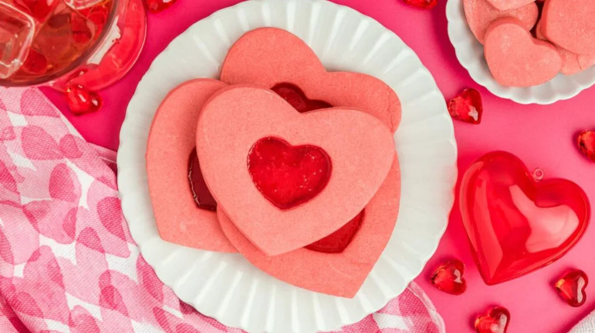 Heart-shaped pink cookies with cut-out centers are arranged on a white plate, surrounded by red heart decorations on a pink background.