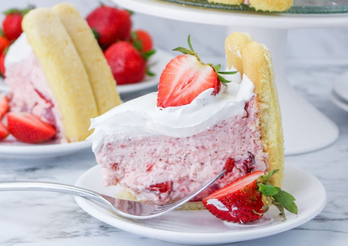 A slice of strawberry mousse cake with whipped cream and a fresh strawberry on top, served on a white plate with a fork and extra strawberries.