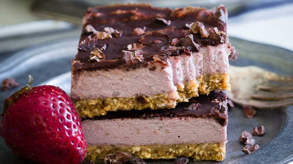Two stacked slices of layered dessert with a nutty crust, pink filling, and chocolate topping, served on a metal plate with a whole strawberry and a fork.
