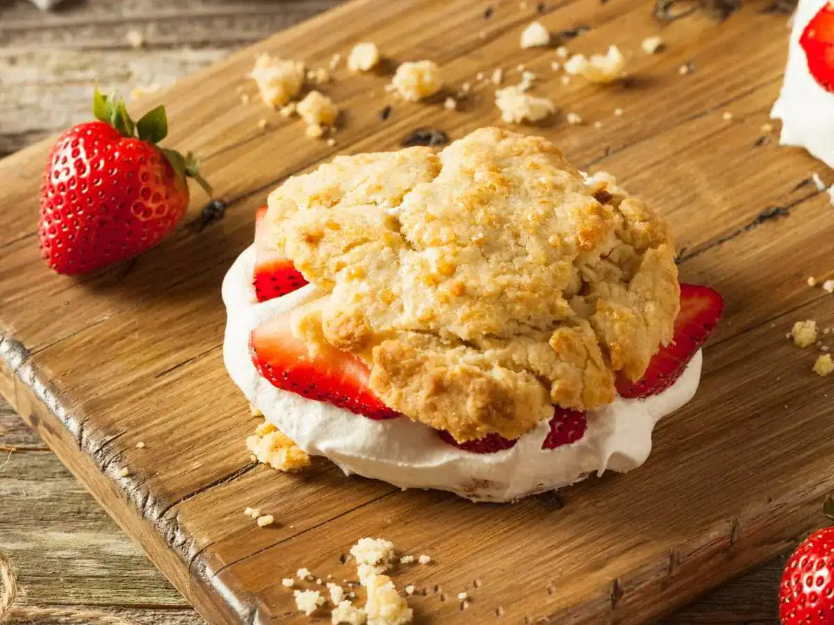 A strawberry shortcake with whipped cream and fresh strawberries sits on a wooden board, surrounded by crumbs and two whole strawberries.