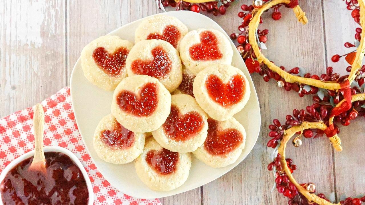 A plate of round cookies with heart-shaped centers filled with red jam, surrounded by red berry and twig decorations on a wooden surface.