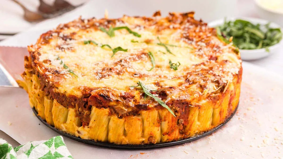A baked pasta pie with rigatoni noodles standing upright, topped with melted cheese and garnished with fresh basil.