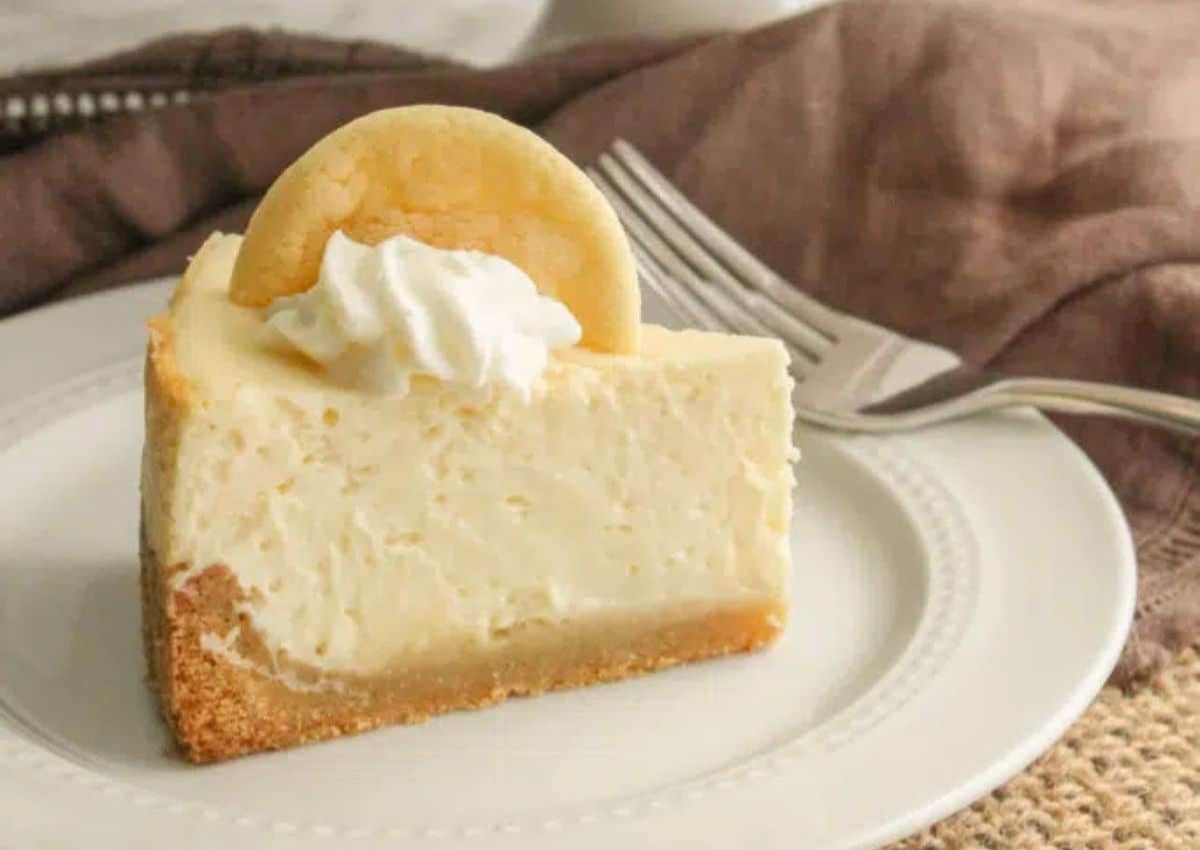 A slice of cheesecake on a white plate, topped with a dollop of whipped cream and a vanilla wafer, with a fork in the background.