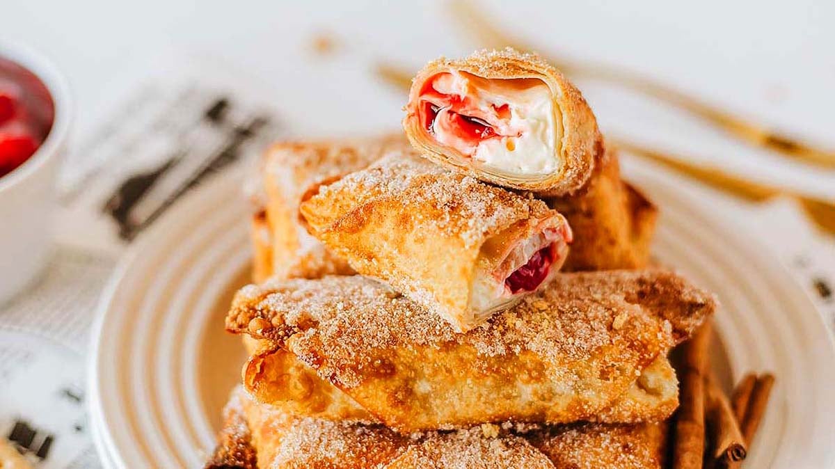 A plate of golden-brown dessert chimichangas filled with cream and cherries, some cut open to show the filling, sprinkled with sugar and cinnamon.
