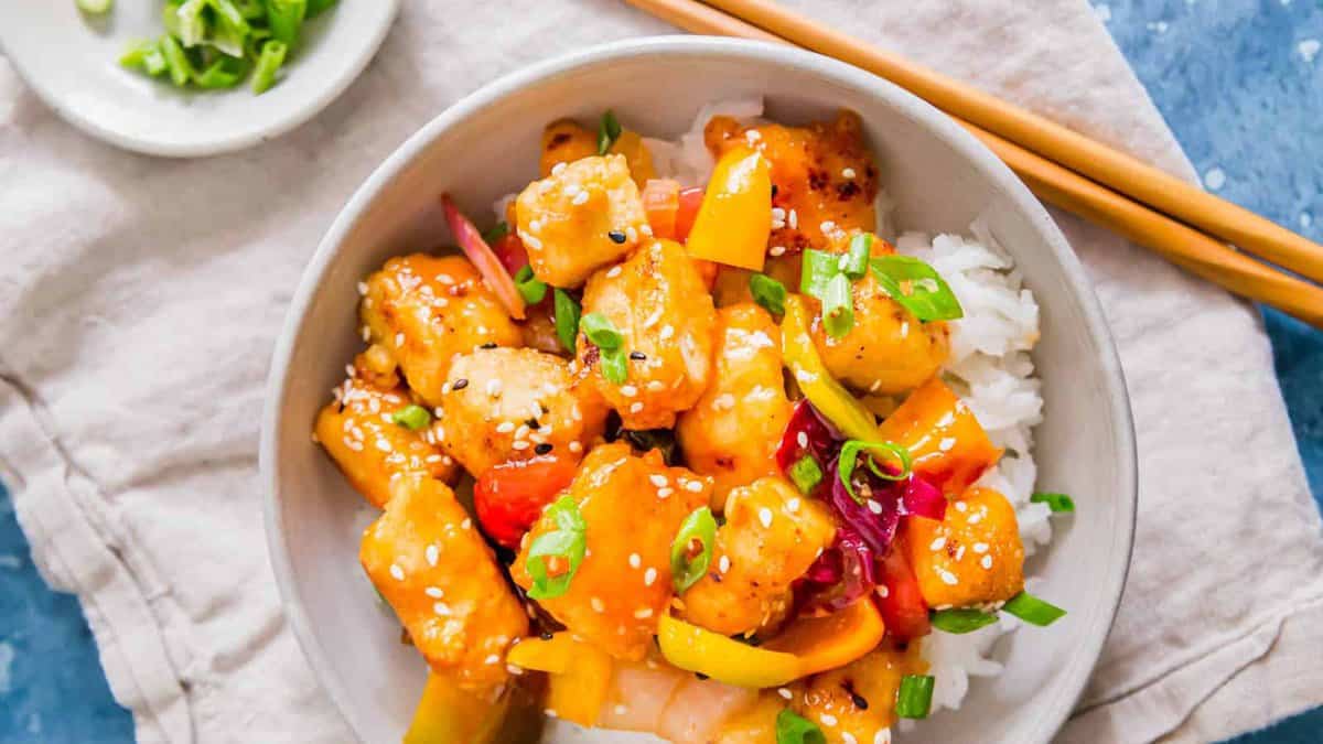 A bowl of sweet and sour chicken with bell peppers, onions, and sesame seeds served over white rice, with chopsticks on the side.