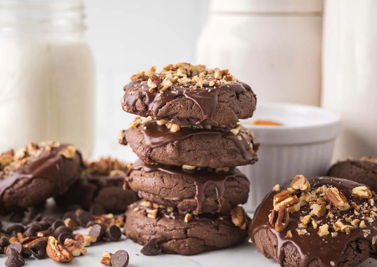 A stack of chocolate cookies topped with chopped nuts and drizzled chocolate, with more cookies, chocolate chips, and jars in the background.