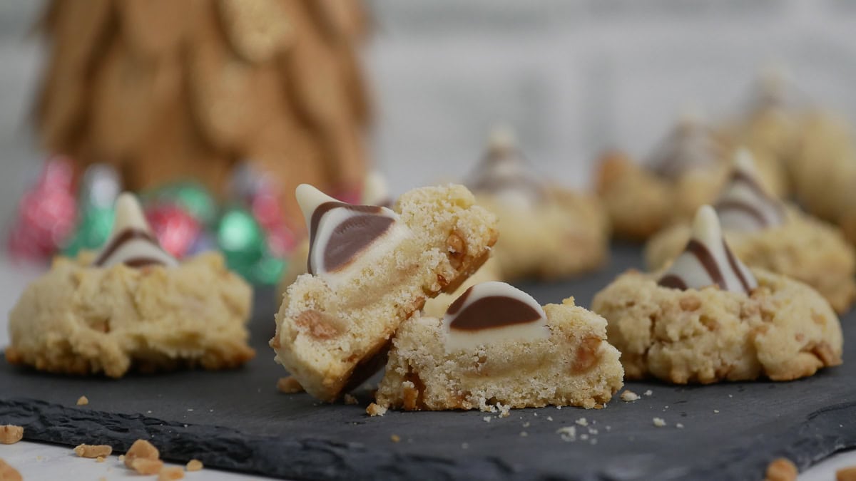 A close-up of crumbly cookies topped with swirled chocolate kisses, displayed on a dark tray with decorative holiday wrappers in the background.