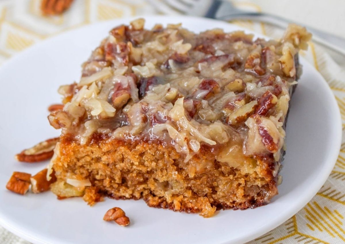 A slice of cake topped with a mixture of chopped pecans and shredded coconut sits on a white plate.
