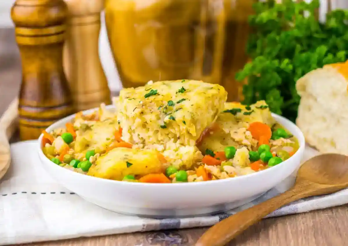 A bowl of chicken pot pie casserole topped with biscuit pieces, mixed with peas, carrots, and sauce, garnished with parsley. A wooden spoon, bread, and seasonings are nearby.