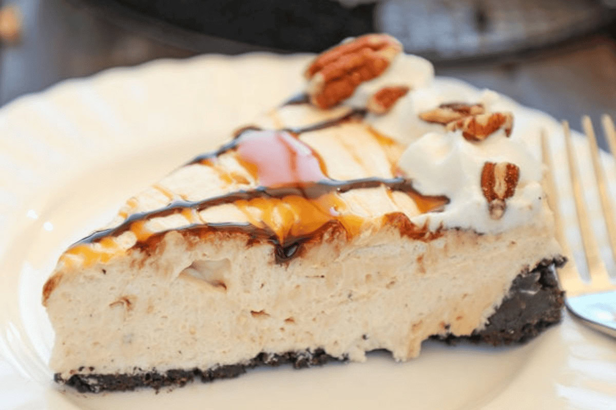 A slice of creamy cheesecake with a chocolate crust, topped with whipped cream, pecans, and drizzles of chocolate and caramel sauce on a white plate.
