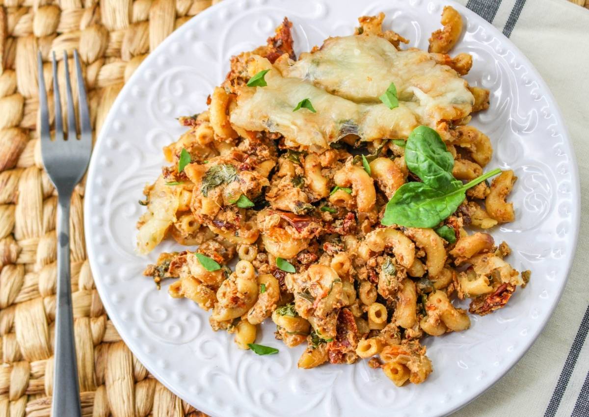 A white plate with baked macaroni mixed with herbs, spinach, and sun-dried tomatoes, topped with melted cheese, and garnished with fresh spinach leaves. A fork rests beside the plate.