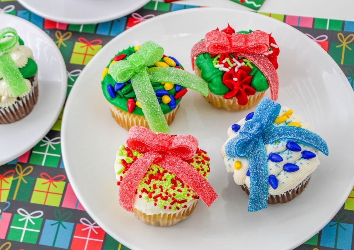 Four cupcakes decorated with colorful frosting, sprinkles, and candy bows arranged on a white plate, set on a gift-wrapped patterned table.