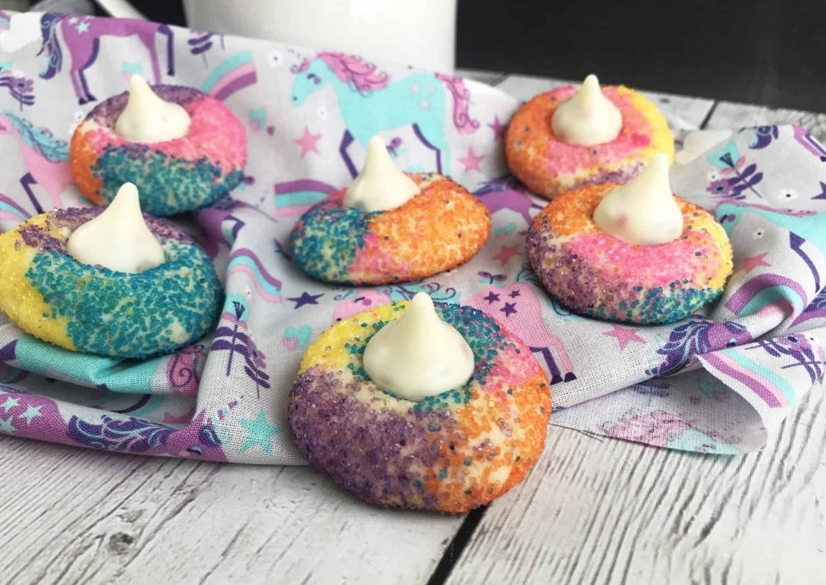 Colorful sugar cookies with white chocolate kisses on top, arranged on a unicorn-patterned cloth on a wooden surface.